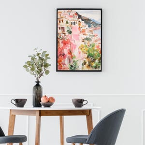 May include: A watercolor painting of a colorful Italian village with pink buildings, green trees, and a blue ocean in the background. The painting is framed in a black frame.