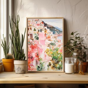 May include: A watercolor painting of a colorful Italian village with pink buildings, green trees, and a blue sea in the background. The painting is framed in a light wood frame.