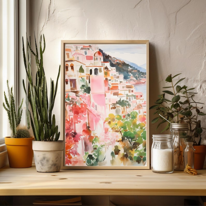 May include: A watercolor painting of a colorful Italian village with pink buildings, green trees, and a blue sea in the background. The painting is framed in a light wood frame.