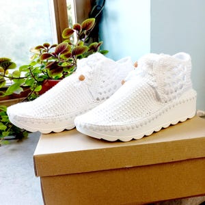May include: A pair of white crochet sneakers with a textured design and a scalloped sole. The sneakers have a high-top design with decorative buttons. They are displayed on a brown cardboard box.