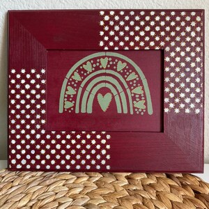 May include: A burgundy wood frame with a white polka dot pattern on the top and left side. The frame contains a green painted rainbow with white hearts.