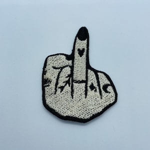 May include: White embroidered patch of a middle finger with black stitching and a heart and spade tattoo on the finger.