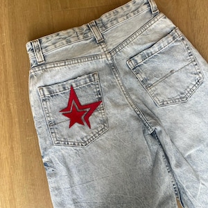 May include: Light blue acid-wash denim jeans featuring a high-waisted design with belt loops and two back pockets. A prominent red star-shaped patch with a dark outline is stitched onto the left back pocket, adding a distinct visual detail.