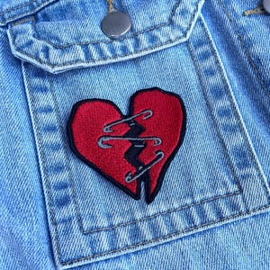May include: A light blue denim jacket pocket featuring an embroidered patch of a red heart split in two. The heart halves are held together by three silver-coloured safety pin designs stitched in black thread, creating a jagged seam effect.