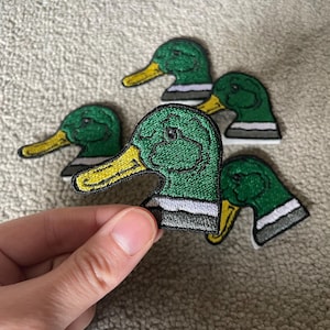 May include: Embroidered duck head patches in green, yellow, white, and black. The patches feature a detailed design of a duck's head and neck. The patches are likely for clothing or accessories.