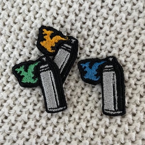 May include: Embroidered patches featuring spray paint cans with colorful flames in yellow, green, and blue. The patches have a black border and are set against a textured, off-white background.
