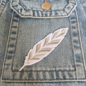 Könnte beinhalten: Eine hellblaue Jeansjackentasche mit einem silbernen, metallisch bestickten Federaufnäher. Der Aufnäher ist mittig auf der rechteckigen Taschenklappe positioniert, die einen einzelnen silberfarbenen Metallknopf am oberen Rand aufweist.