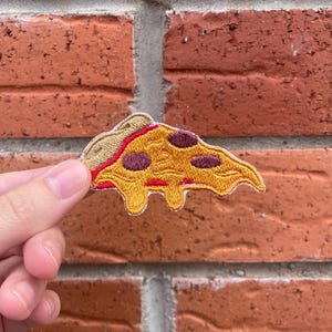 Op de afbeelding: Een geborduurde patch in de vorm van een pizzapunt met een goudgele korst, een laag rode tomatensaus en donkerbruine ronde pepperoni-toppings. De kaas is afgebeeld als smeltend in drie afzonderlijke druppels onder de punt.