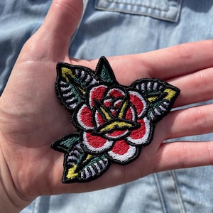 Könnte beinhalten: Gestickter Aufnäher mit einer klassischen roten Rose, grünen Blättern und gelben Akzenten. Der Aufnäher hat einen schwarzen Rand und ist wahrscheinlich zum Aufbügeln oder Aufnähen auf Stoff gedacht. Das Design erinnert an traditionelle Tattoo-Kunst.
