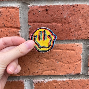 May include: A colourful, embroidered patch featuring a yellow smiley face with a distorted, melting design. The smiley face has black outlines and red, blue, and yellow accents. The patch is held against a red brick wall.
