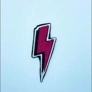 May include: A pink lightning bolt iron-on patch with a white outline and black stitching.