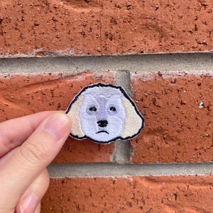 May include: Embroidered patch featuring a sad-faced dog with a light purple face, black nose and eyes, and tan ears. The patch is outlined in black and is held against a red brick wall.