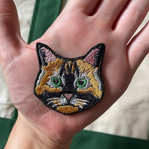 May include: Embroidered cat face patch with a black border. The patch shows a calico cat with green eyes, orange, yellow, and black fur, and a pink nose. The patch is held in a hand.