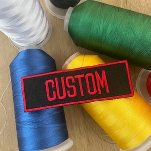 Può includere: Una toppa nera con bordi rossi e la parola "CUSTOM" ricamata in rosso. La toppa è circondata da rocchetti di filo di vari colori, tra cui bianco, nero, verde, blu e giallo. I fili sono avvolti su rocchetti di plastica bianca.