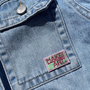 May include: A light blue denim jacket featuring a chest pocket with a silver-toned metal button. A rectangular embroidered patch is sewn onto the pocket, displaying the text "MAKE ART." in dark pink letters on a light green background.