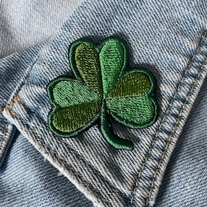 May include: A green embroidered shamrock patch on a light blue denim jacket collar. The shamrock has three leaves with varying shades of green and a dark green outline. The denim jacket has visible stitching.