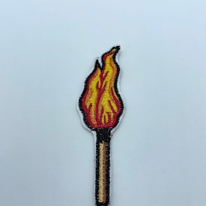 Puede incluir: Un parche bordado que representa una cerilla con una llama en la parte superior, en rojo y amarillo.