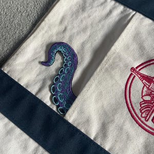 May include: Cream canvas tote bag with navy blue straps and a pocket. A purple and teal octopus tentacle patch is sewn onto the pocket. A red and white embroidered design is visible on the bag.
