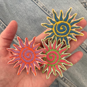 Può includere: Tre toppe ricamate a forma di sole con centri a spirale e punte radianti. Una toppa ha un centro blu con raggi gialli, una ha un centro viola con raggi arancioni e una ha un centro verde con raggi rosa, tutte con bordi bianchi.