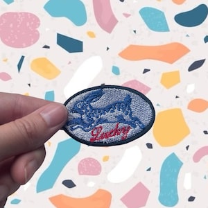 Op de afbeelding: Een ovaalvormige patch met een blauw konijnontwerp en het woord "Lucky" in rood script. De patch heeft een zwarte rand. De achtergrond is een kleurrijk, abstract patroon.
