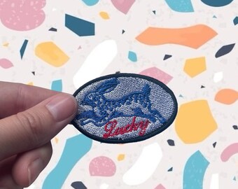 May include: An oval-shaped patch with a blue rabbit design and the word "Lucky" in red script. The patch has a black border and is held against a colorful, abstract background.