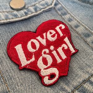 May include: A red heart-shaped patch with the words "Lover Girl" embroidered in white. The patch is sewn onto a light blue denim jacket, with a silver button visible at the top.