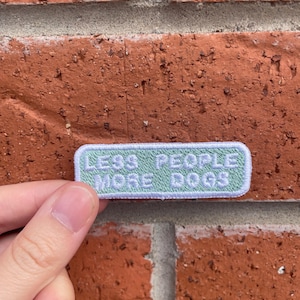 Może przedstawiać: Prostokątna naszywka z napisem "LESS PEOPLE MORE DOGS" w białych literach na jasnym zielonym tle. Naszywka jest trzymana na tle czerwonej ceglanej ściany.
