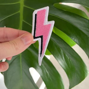 Peut inclure: Patch thermocollant en forme d'éclair rose avec un contour noir. Le patch est sur un fond de feuille verte.