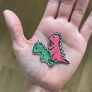 May include: Two embroidered dinosaur patches. One green dinosaur is lying down, and a pink dinosaur is standing on top of it. Both patches have black outlines and white stitching.