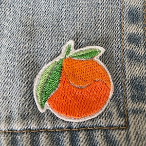 Op de afbeelding: Geborduurde patch met een oranje vrucht met groene bladeren. De oranje vrucht heeft een heldere oranje kleur met een witte rand. De patch is genaaid op een blauwe spijkerstof.
