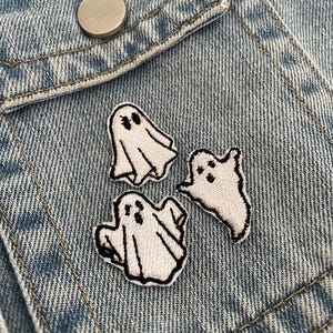 Op de afbeelding: Drie witte spookpatches met zwarte geborduurde contouren zijn bevestigd op een lichtblauwe spijkerjas. De patches variëren in vorm en grootte, elk met twee kleine zwarte stippen voor ogen. Een zilveren knoop is zichtbaar.