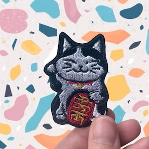 May include: Embroidered patch with a Maneki-neko cat design. The patch shows a grey and white cat with a black outline, a red collar, and a gold coin. The cat is in a welcoming pose, with one paw raised.