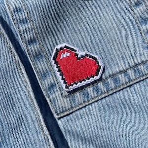 Peut inclure: Gros plan d'une poche de veste en jean bleu clair ornée d'un écusson cœur rouge pixélisé. Le cœur est bordé de noir et de blanc. La veste présente des coutures et un design classique.