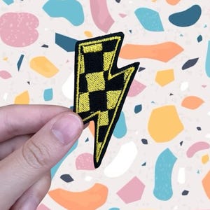 May include: An embroidered lightning bolt patch in black and yellow, featuring a checkered design. The patch is held against a colourful terrazzo background. This patch can be ironed onto clothing or accessories.