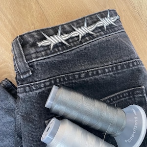 Könnte beinhalten: Eine schwarze Jeans aus Denim mit einem weißen, gestickten Stacheldrahtmuster am Bund. Auf der Jeans liegen zwei Garnrollen, eine in metallischem Silber und die andere in einem helleren, glänzenden Weißton.