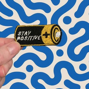 Può includere: Toppa ricamata a forma di batteria con la scritta bianca "STAY POSITIVE". La batteria è nera e gialla, con un dettaglio a forma di segno più. Lo sfondo è color crema con linee e cerchi blu.
