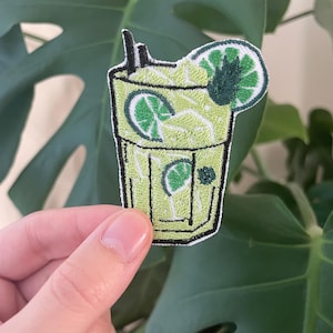 May include: Embroidered patch featuring a green cocktail with lime slices and a straw. The patch has a black border and is held against a backdrop of green leaves. The cocktail design includes a lime wedge garnish.