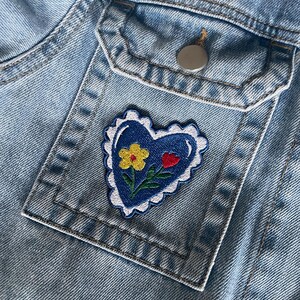 May include: Close-up of a light blue denim jacket with a pocket. A blue heart-shaped patch with a white scalloped border is sewn onto the pocket. The patch features a yellow flower, a red heart, and green leaves.