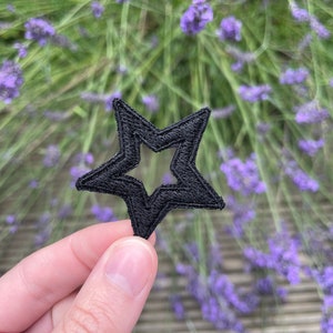 May include: A black star-shaped patch with a smaller star cut out in the centre. The patch is held in a hand against a blurred background of purple lavender flowers.