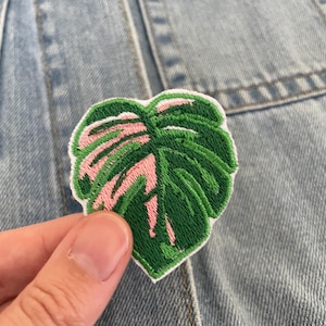May include: Embroidered patch featuring a green and pink monstera leaf design. The patch has a white border and is held against a light blue denim jacket. The leaf design is detailed with green and pink thread.