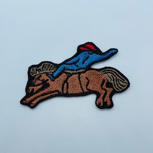 May include: Embroidered patch of a brown horse with a blue and red cowboy riding on its back. The cowboy is wearing a red hat and blue shirt.