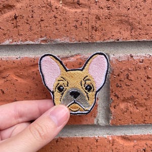 May include: An embroidered patch featuring the head of a French Bulldog with tan fur, large pink-lined ears, black eyes, and a dark muzzle. The patch has a thick black border and is shaped to follow the outline of the dog's head and ears.