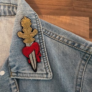 Peut inclure: Veste en jean bleu clair avec un écusson décoratif. L'écusson présente un motif doré orné au-dessus d'un cœur rouge percé d'un poignard argenté. La veste a une fermeture boutonnée.