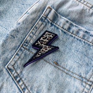 May include: A light blue acid-wash denim fabric featuring a lightning bolt patch. The patch has a leopard print centre with black and purple embroidered borders, securely attached to the back pocket area of the denim garment.