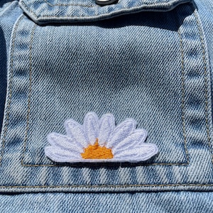 May include: A light blue denim jacket with a white daisy flower patch with an orange centre in the pocket.