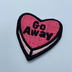Può includere: Una toppa ricamata a forma di cuore rosa con le parole "Go Away" in lettere nere. La toppa ha un bordo nero e un contorno rosa chiaro, che ricorda un cuore di caramella.