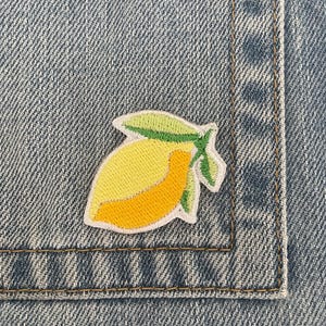 Può includere: Toppa ricamata a forma di limone con frutta gialla e arancione e foglie verdi, cucita su una tasca di jeans. La toppa ha un bordo bianco ed è cucita con filo giallo, arancione e verde.