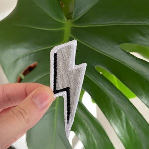 May include: A white lightning bolt iron-on patch with a black outline. The patch is held in a hand against a green leafy background.