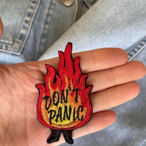 May include: Embroidered patch in the shape of flames with the words "DON'T PANIC" in yellow. The patch has a black border and small black feet. The patch is held in a hand, with a denim jacket in the background.