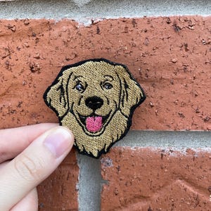 Könnte beinhalten: Ein gestickter Aufnäher mit dem Kopf eines Golden Retrievers, der beiges Fell, schwarze Augen, eine schwarze Nase und eine leuchtend rosa Zunge zeigt. Der Aufnäher hat einen schwarzen gestickten Rand und folgt der Form des Kopfes und der Ohren.